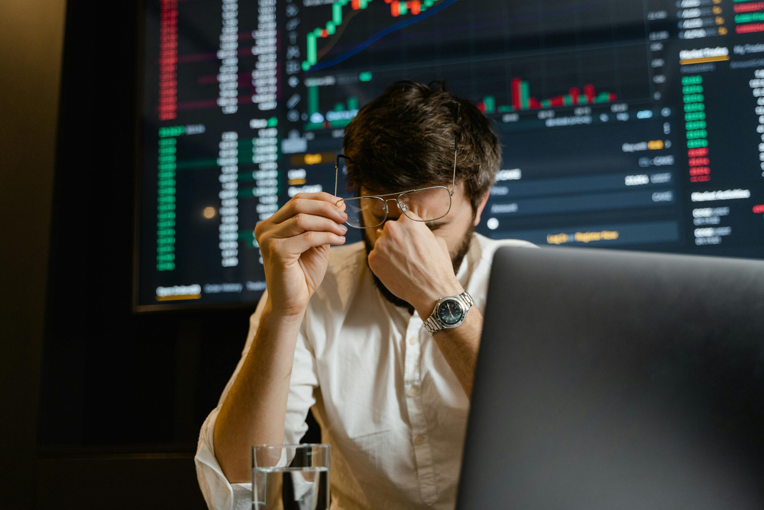 Man rubbing his eyes in front of a computer with stock data, suggesting stress from overtime work